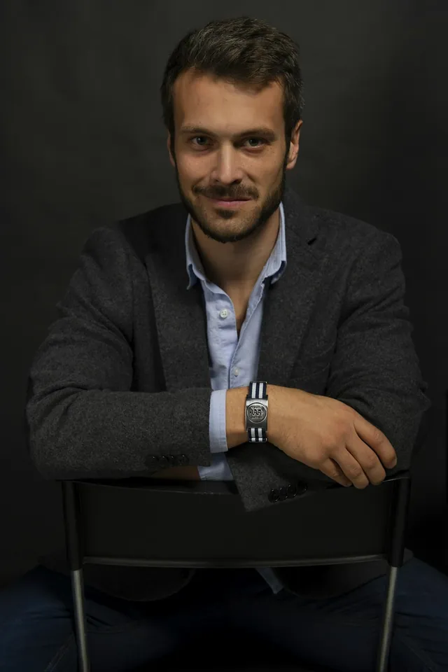 A man in a charcoal jacket seated against a dark studio backdrop (illustrative)